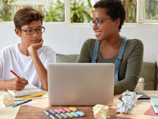 A teenager studying with a smiling tutor using a laptop at a cluttered desk.