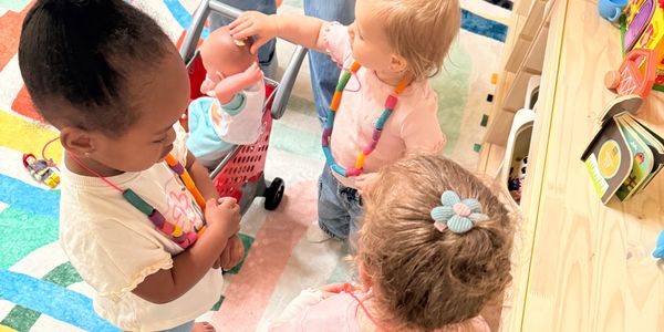 Three toddlers playing with dolls and toys in a colorful playroom.