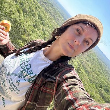 Woman taking a selfie with a scenic forest backdrop on a sunny day.