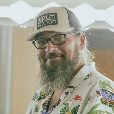 Bearded man wearing glasses and a tropical shirt smiles warmly.