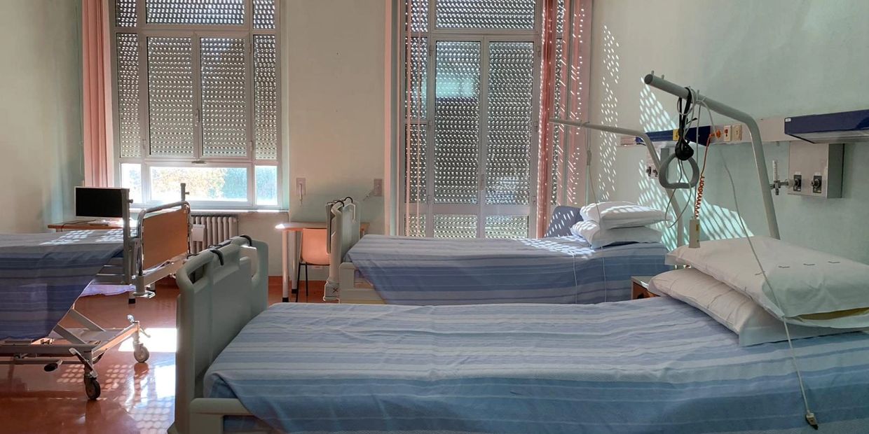 Empty hospital room with three beds and sunlight through windows.