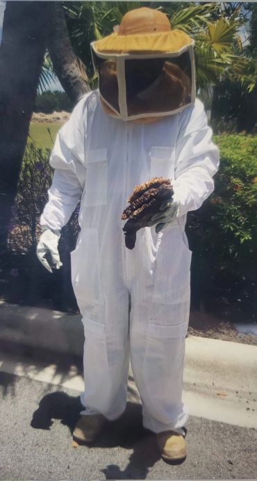 Person in a beekeeping suit holding a honeycomb outdoors.