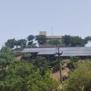 Solar panels installed on a hillside with trees and a building in the background.