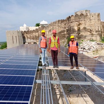 Three workers in safety gear standing on solar panel installation near old fort.