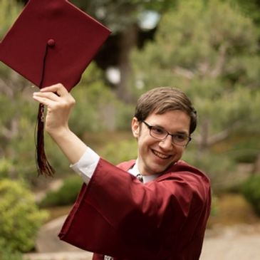 A white man smiling. He is wearing a red graduation cap and gown. He has short brown hair.