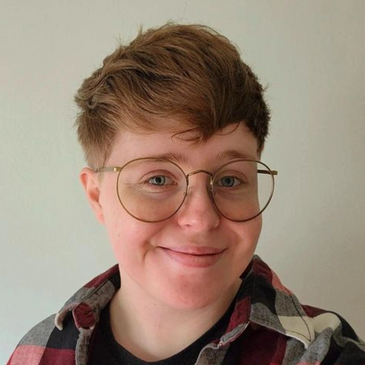 A white, non-binary person. They are smiling, wearing a red flannel shirt, and round gold glasses.