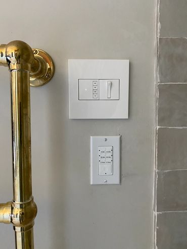 Two white timer switches mounted on a beige wall next to brass pipe and tiled wall.