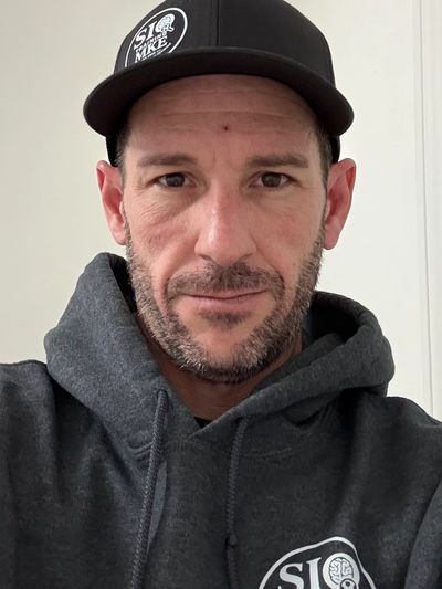 Man wearing a black cap and dark hoodie with logos, looking directly at the camera.