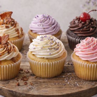 Six assorted cupcakes with colorful frosting on a wooden board.