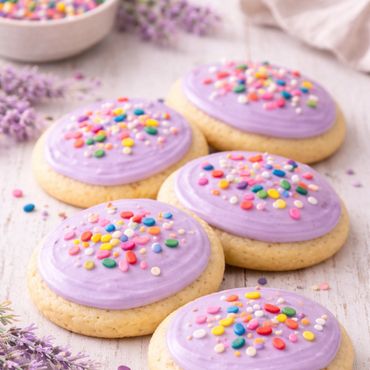 Sugar cookies with purple frosting and colorful sprinkles on a white wooden surface.