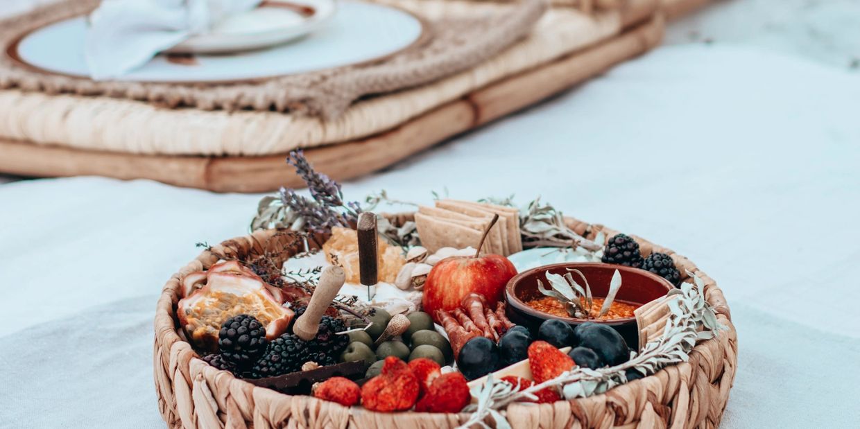 A round cheese and charcuterie platter is set against a white linen background. A beach picnic