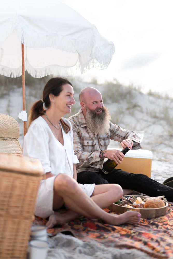 A man opens a wine bottle while a woman sits beside him on a picnic rug smiling. at the beach