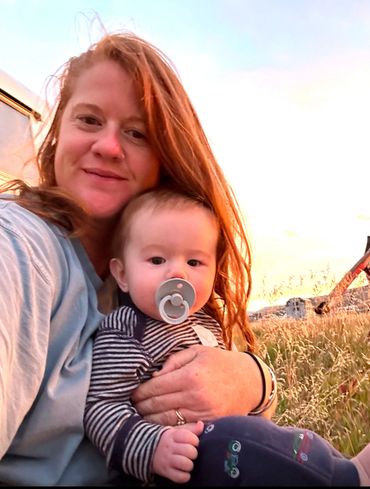A woman with red hair holding a baby with a pacifier outdoors at sunset.
