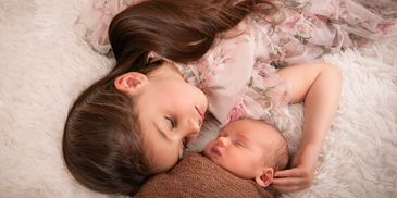 A young girl lovingly cuddles a sleeping newborn baby on a soft blanket.