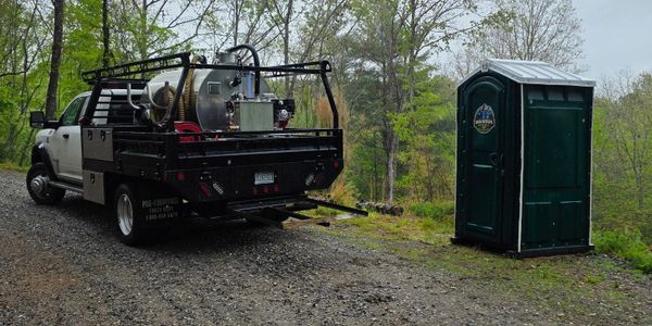 Truck with equipment parked near a portable toilet in a wooded area.