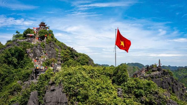 Vietnamese flag atop a rocky mountain with visitors and temple structures.