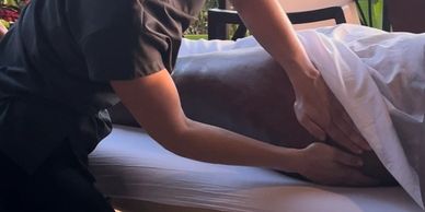 Woman in a grey shirt and black yoga pants massaging a woman on a massage table with white sheets