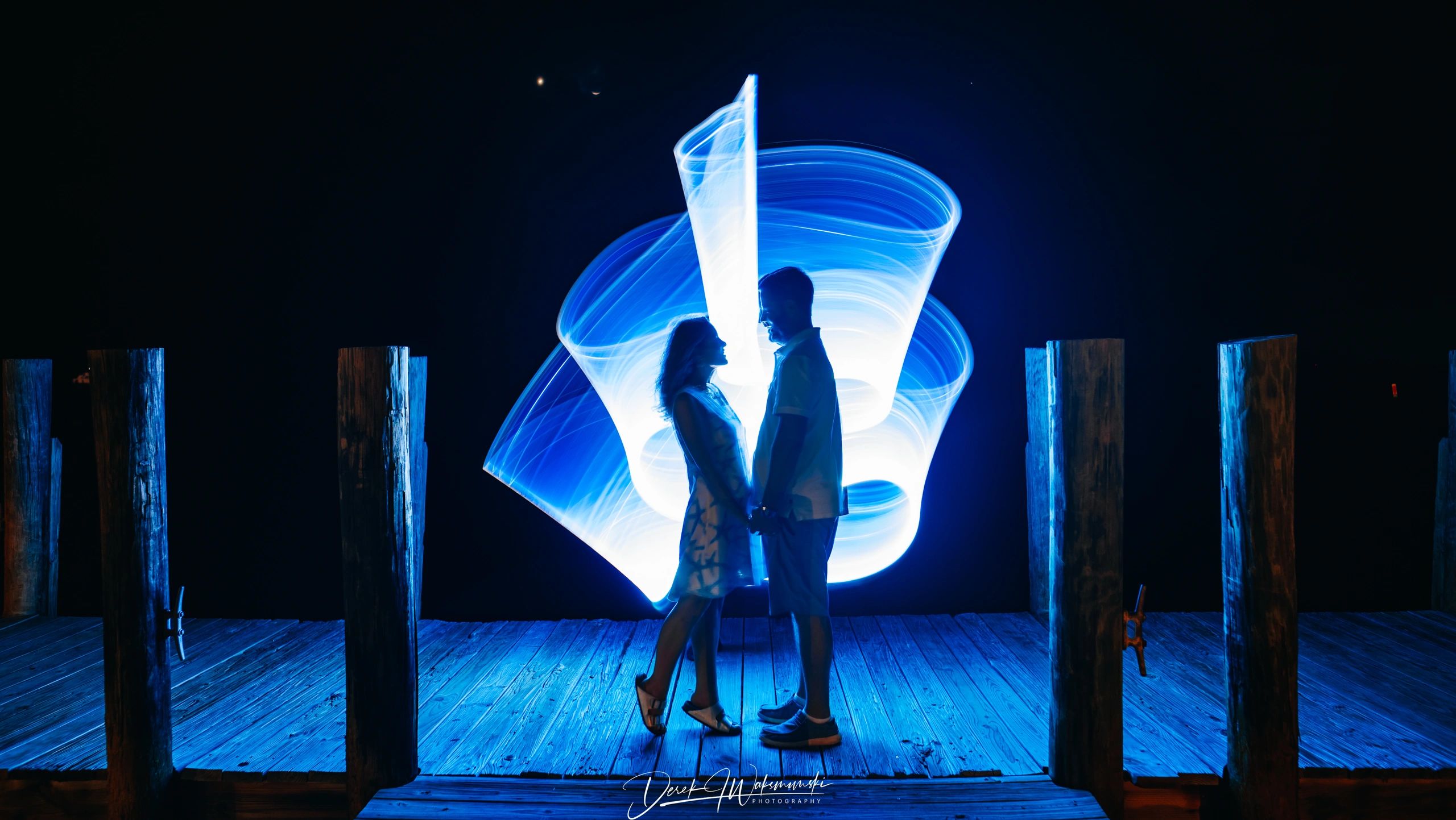 Couple holding hands with blue light trails at night on a wooden dock.