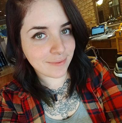 Woman with dark hair, facial piercings, and chest tattoo wearing a red plaid shirt.