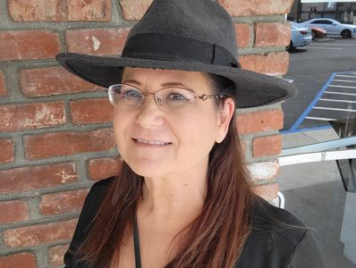 Smiling woman wearing glasses and a black hat standing by a brick wall.