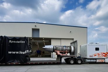 Large jet engine mounted on a flatbed trailer outside an industrial building.