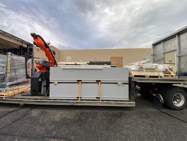 Flatbed truck carrying industrial equipment and a robotic arm under a cloudy sky.