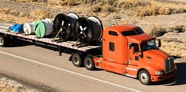 Orange semi-truck hauling large cable reels on a flatbed trailer on a desert highway.