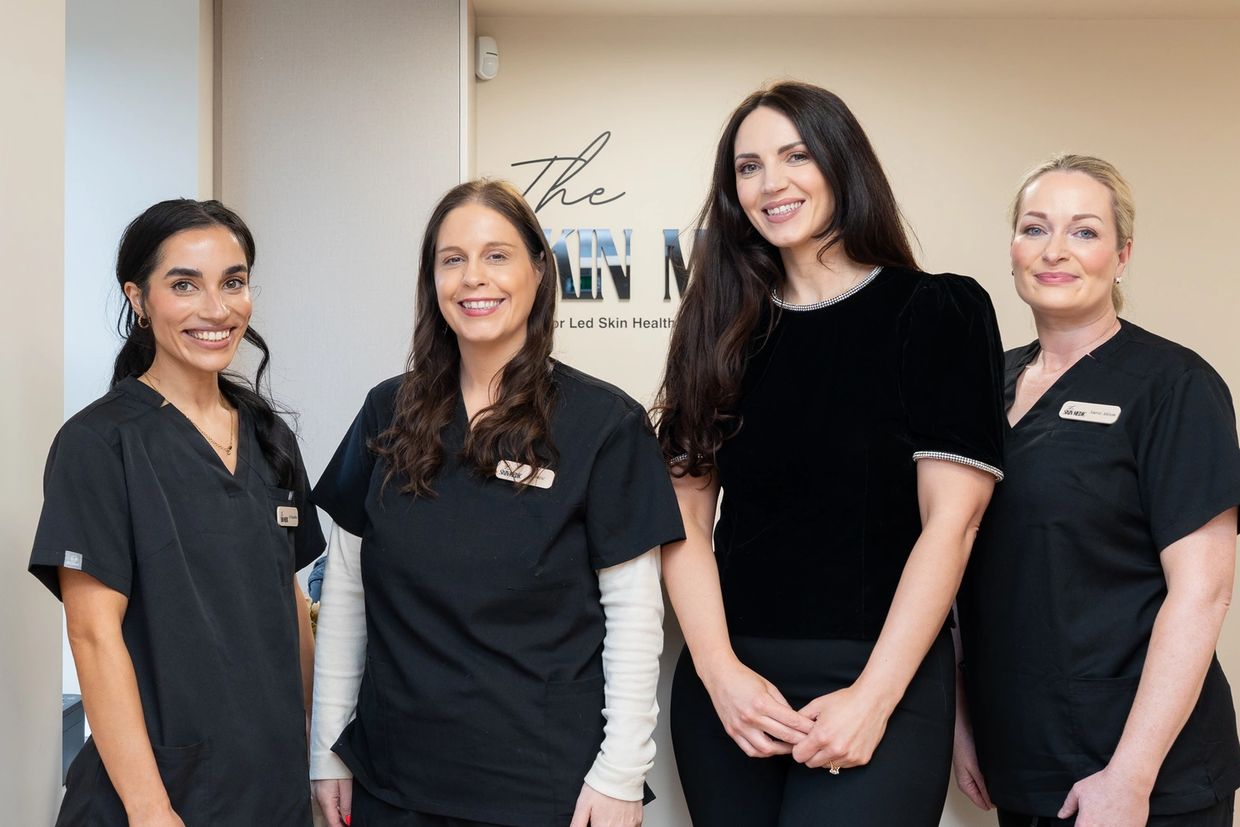 Four smiling women, three in black scrubs and one in a black top, standing in a clinic.