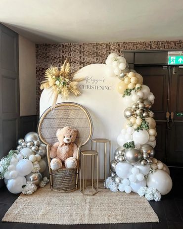 Elegant christening setup with balloons, a teddy bear, and floral decor.