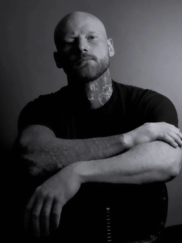 Black and white portrait of a tattooed bald man with a beard, arms crossed.