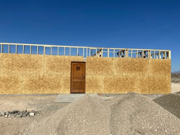 Wooden construction wall with a door and building framework behind it under a clear blue sky.