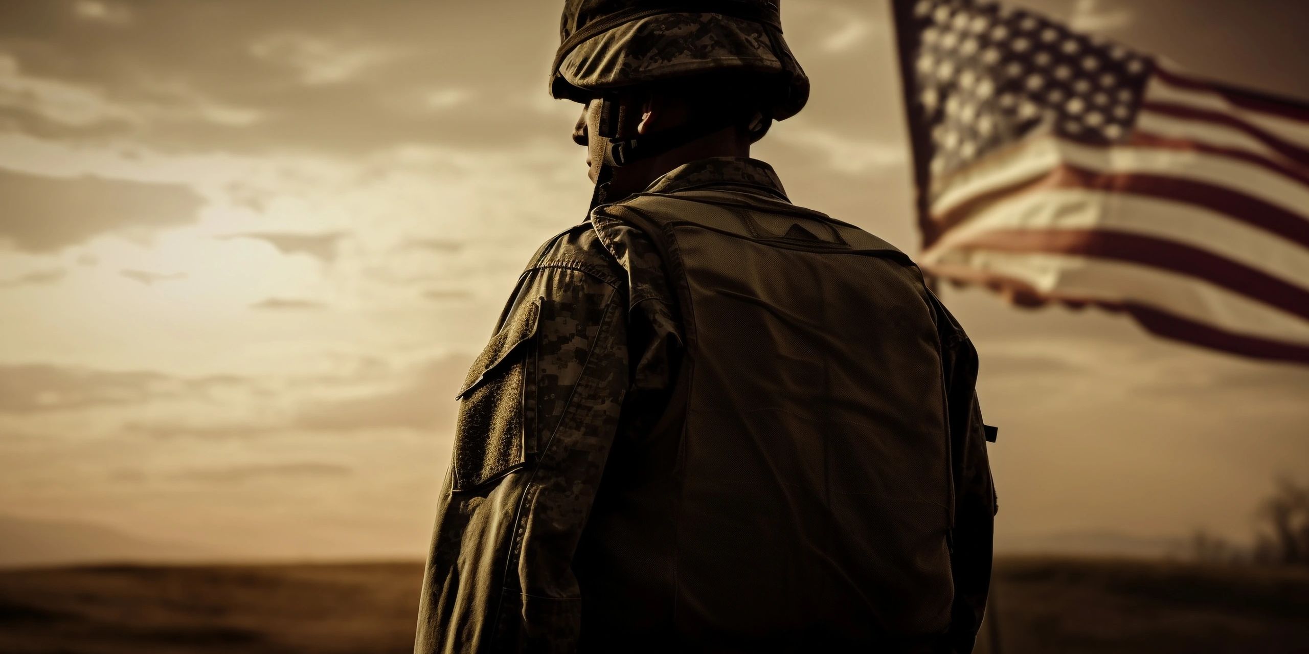 Soldier in military gear stands with the American flag at sunset.