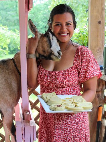 A smiling woman in a red dress holds a plate of goat milk soaps next to a goat.
