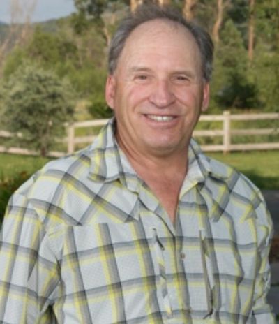 Smiling man in a plaid shirt outdoors with greenery and a fence in the background.