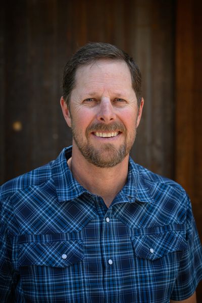 Smiling man in a blue plaid shirt against a wooden background.