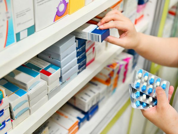 A person's hands reach for a box on a shelf while holding blister packs of pills.