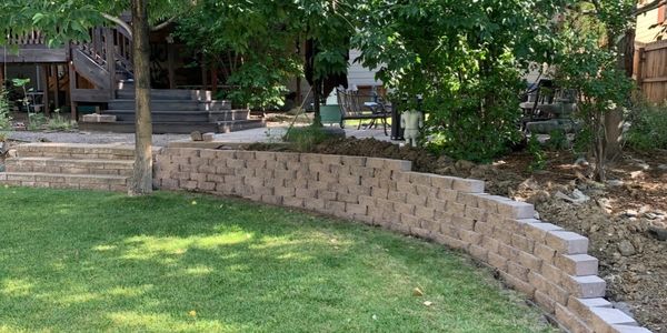 A curved stone retaining wall with staggered height is surrounded by trees and grass in a backyard.