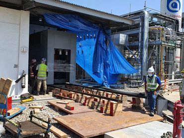 Workers in safety gear at an industrial construction site with equipment and blue tarp.