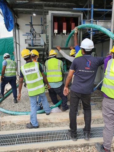 Construction workers handling a large green hose near electrical equipment.
