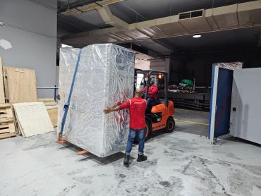 Workers move a large wrapped item using a forklift in a warehouse.