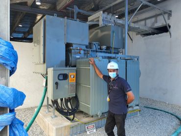 Technician in safety gear standing beside a large electrical transformer.