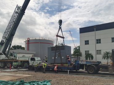 Workers use a crane to lift a large industrial unit onto a flatbed trailer outdoors.