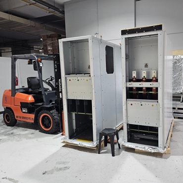 Orange forklift parked next to two large industrial electrical cabinets in a warehouse.
