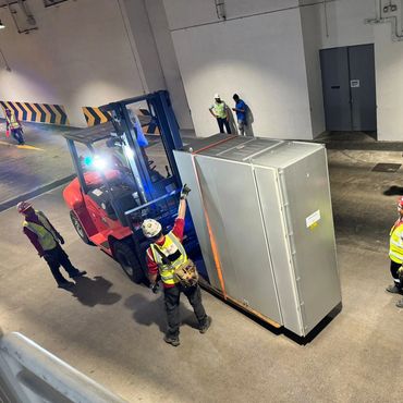Workers with safety gear operating forklift to move large equipment indoors.