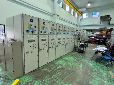 Industrial electrical control panels in a spacious room with high windows.