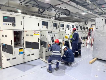 Workers in safety gear maintaining electrical control panels in an industrial setting.