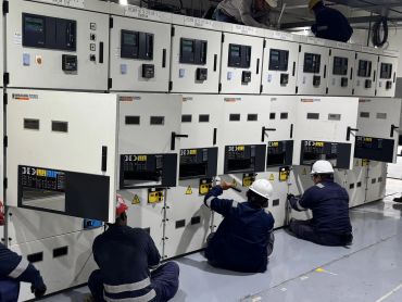 Technicians working on electrical panels in a control room.