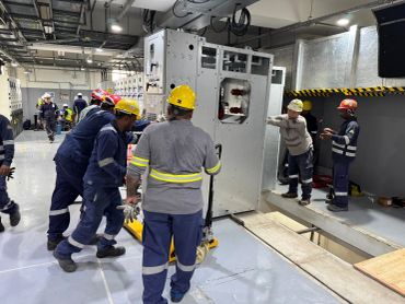 Workers in safety gear install heavy electrical equipment indoors.
