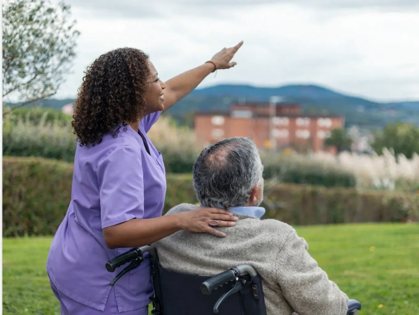 Caregiver pointing out something to an elderly man in a wheelchair outdoors.