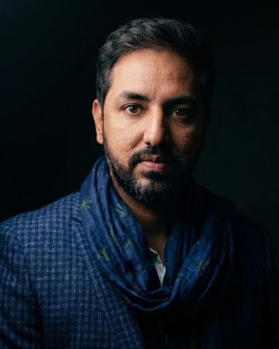 Portrait of a man with a beard wearing a blue checkered blazer and scarf against a dark background.